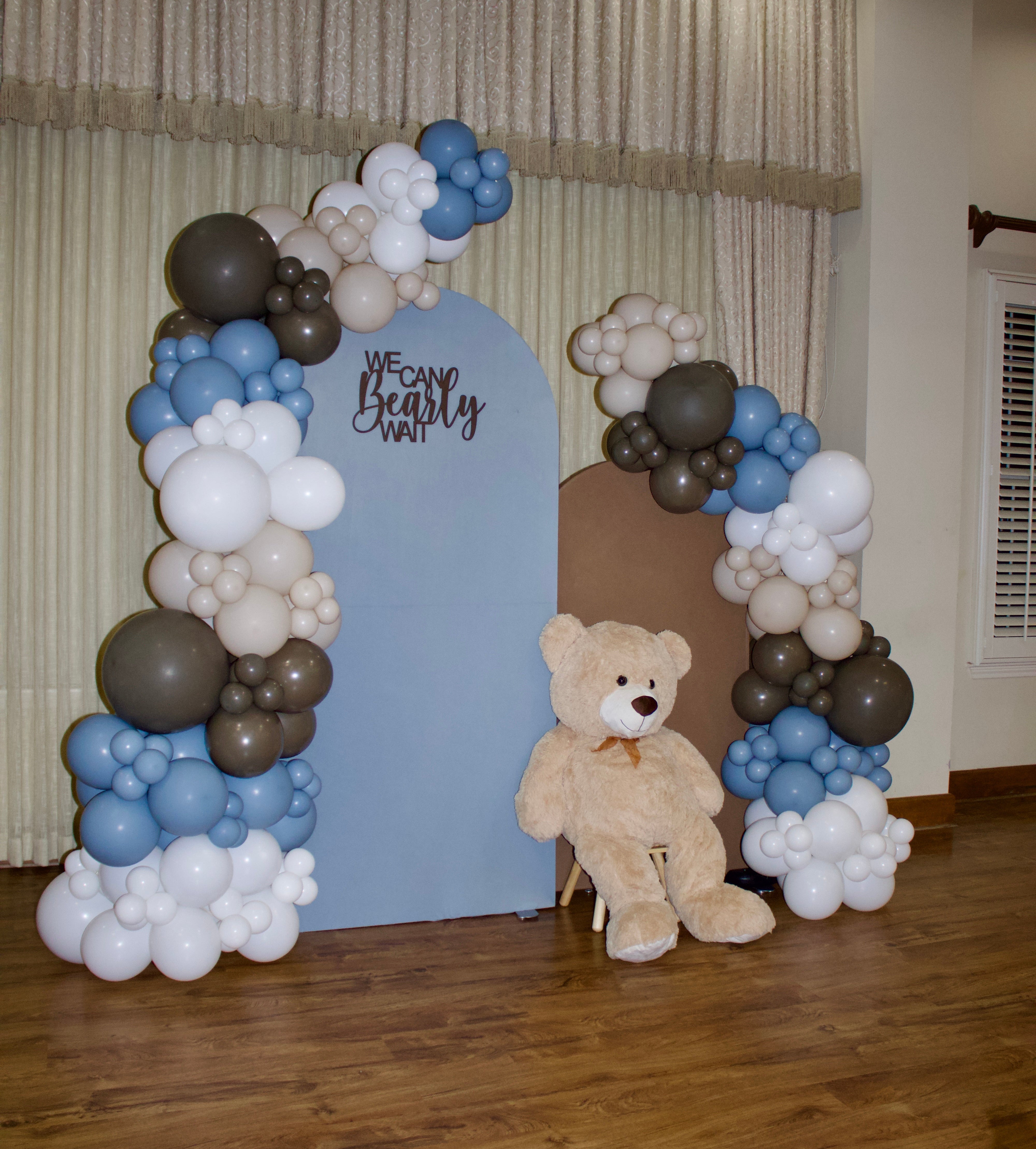 Decorative balloon arch with teddy bear and 'We Can Be Teddy' sign on a wooden floor.